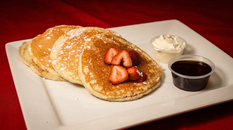 Fluffy pancakes topped with strawberries and powdered sugar, served with syrup and whipped cream.