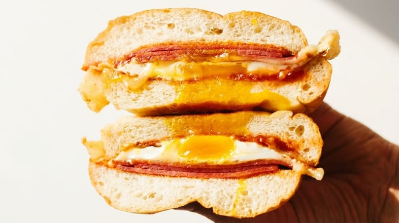 A hand holds a sliced breakfast sandwich with runny egg yolk, cheese, and meat.